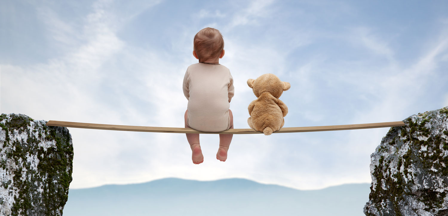 A baby boy and a teddy bear sit on a plank balanced between 2 cliffs, enjoying the view